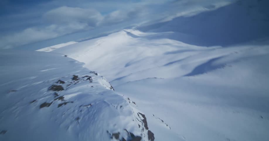 Dynamic FPV aerial footage flies along the edge of a vast, snow-covered mountain range on a clear day