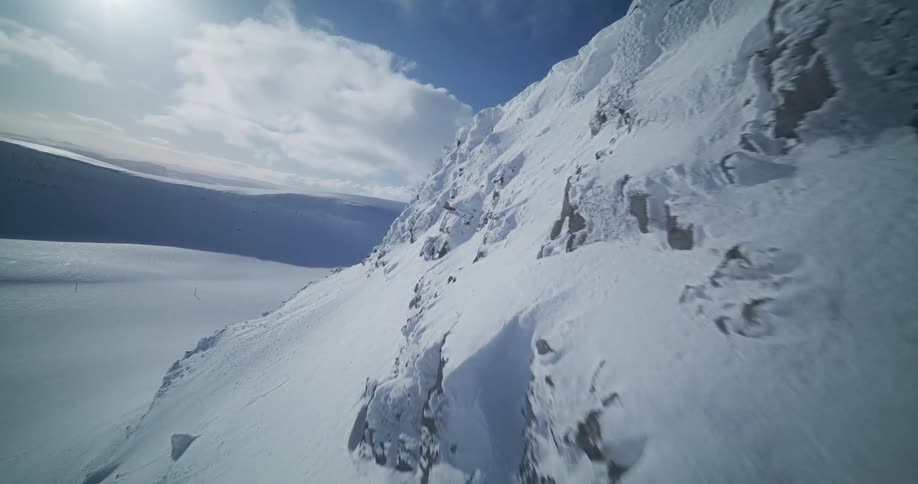 An exhilarating FPV drone flight speeds up a snow-covered mountain slope, closely following the dramatic rocky ridge against a bright sun and blue sky