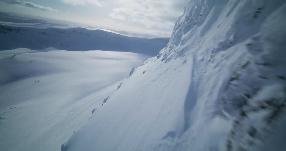 Dynamic FPV drone footage captures a thrilling flight along the edge of a snow-covered mountain cliff in a vast winter landscape