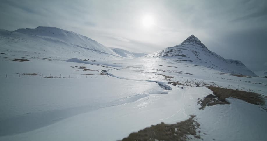 A dynamic aerial shot flies low over a snow-covered landscape, passing traditional fish drying racks towards Iceland's iconic Kirkjufell mountain