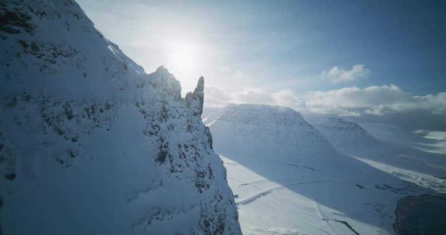 A thrilling FPV drone flight soars along a steep, snow-covered mountain ridge, diving towards a vast, icy valley and fjord below