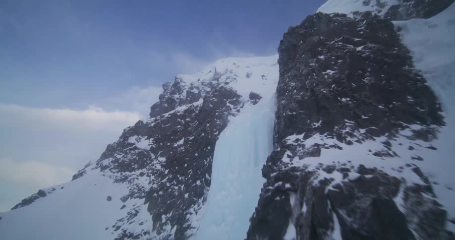 An epic aerial shot ascends a rugged, snow-covered mountain, flying past a frozen icefall to reveal the vast, windy summit