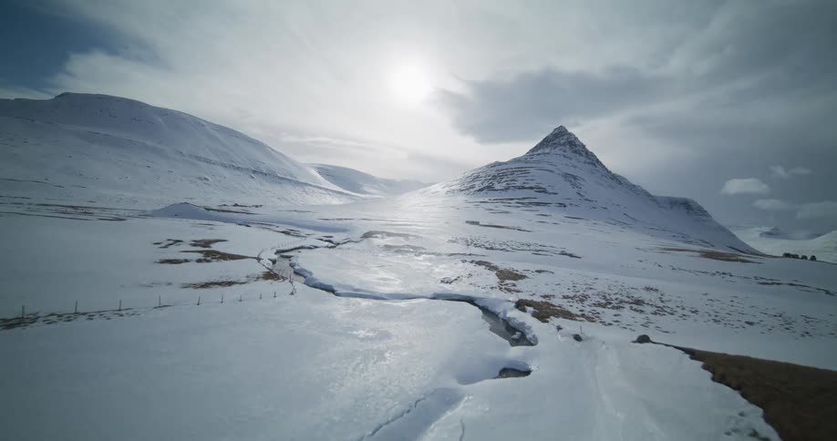 A fast-moving aerial shot glides over a snowy landscape, past wooden structures, towards the iconic Kirkjufell mountain in Iceland