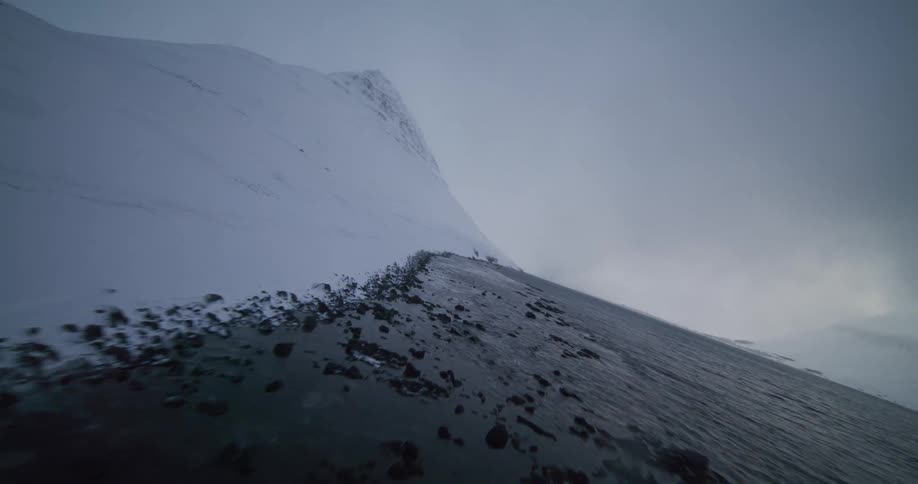 An aerial view follows the rocky, snow-covered coastline of a fjord surrounded by vast mountains in a moody winter landscape