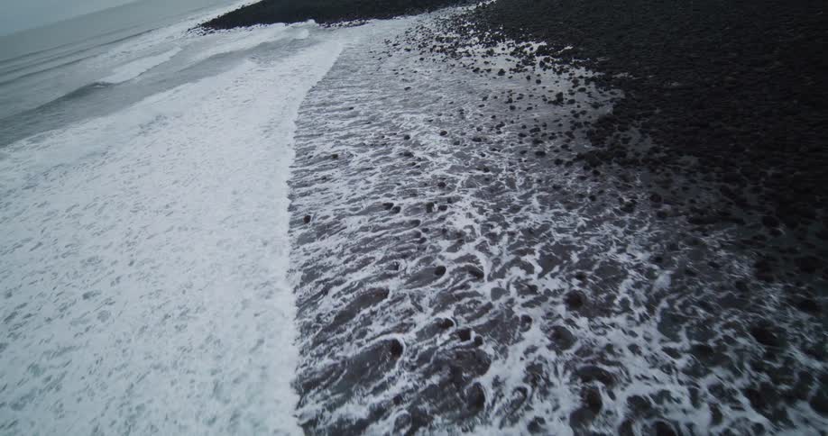 An aerial slow-motion view captures the mesmerizing contrast as white sea foam washes over a dark, rocky coastline