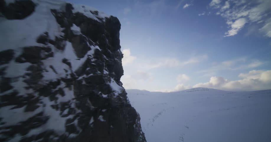 An epic aerial shot soars alongside a rugged, snow-covered mountain cliff revealing a vast winter landscape