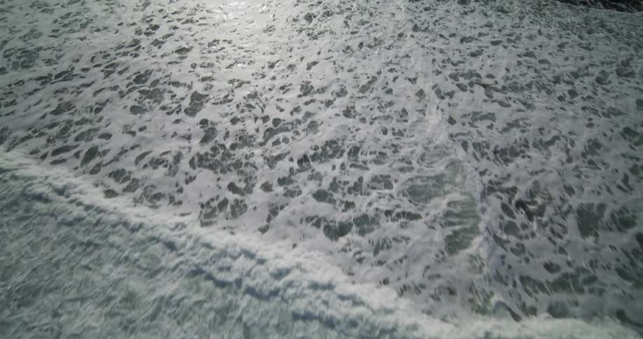 An aerial view slowly moves over the white foamy ocean surf as it rolls onto a dark rocky beach