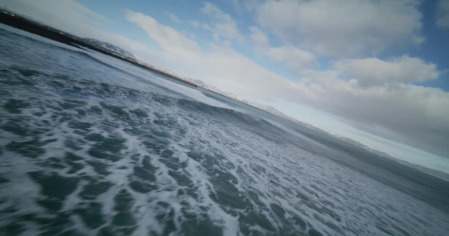 An exhilarating aerial flight passes a stark white tower on a black stone beach and glides low over the ocean waves