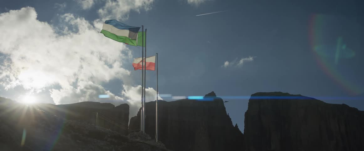 Flags wave proudly on a sun-drenched mountain peak as military jets fly in formation across a dramatic sky