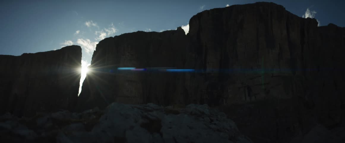 The sun bursts through a crevice in a silhouetted mountain range, casting a dramatic lens flare over the rocky terrain