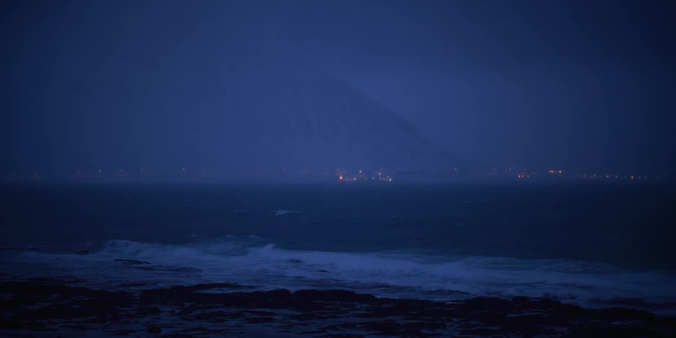 A dramatic shot of a stormy sea at twilight as waves crash on a rocky coast with distant city lights glowing beneath a mountain