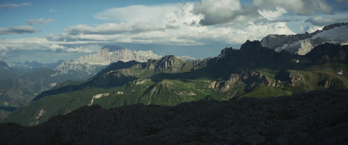 A breathtaking panoramic view pans across a vast mountain range, revealing green valleys and distant snow-capped peaks under a cloudy sky