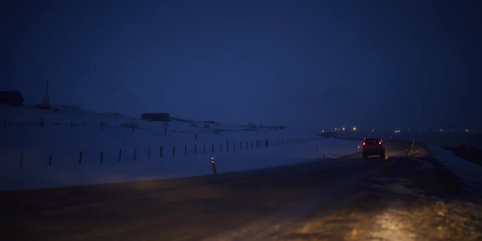 An SUV drives away down a dark, rural road at night through a windy, snow-covered landscape