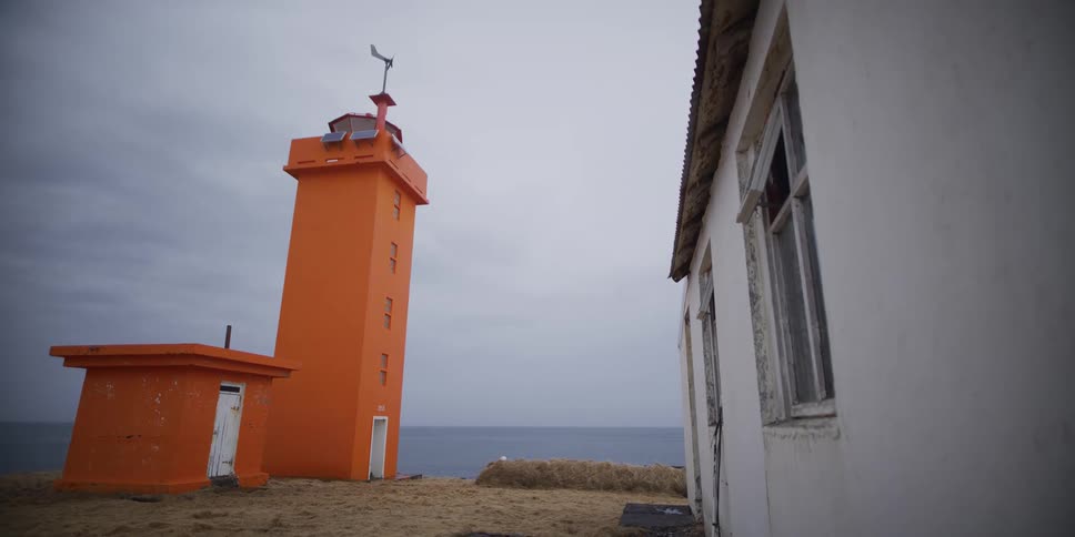 A bright orange lighthouse and a derelict white building stand on a desolate coast overlooking the ocean on a cloudy day