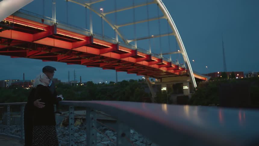Two people hug by a large bridge at sunset
