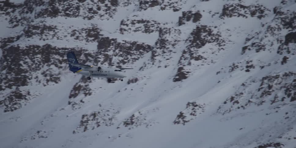 A turboprop airplane on landing approach flies slowly past a rugged, snow-covered mountain
