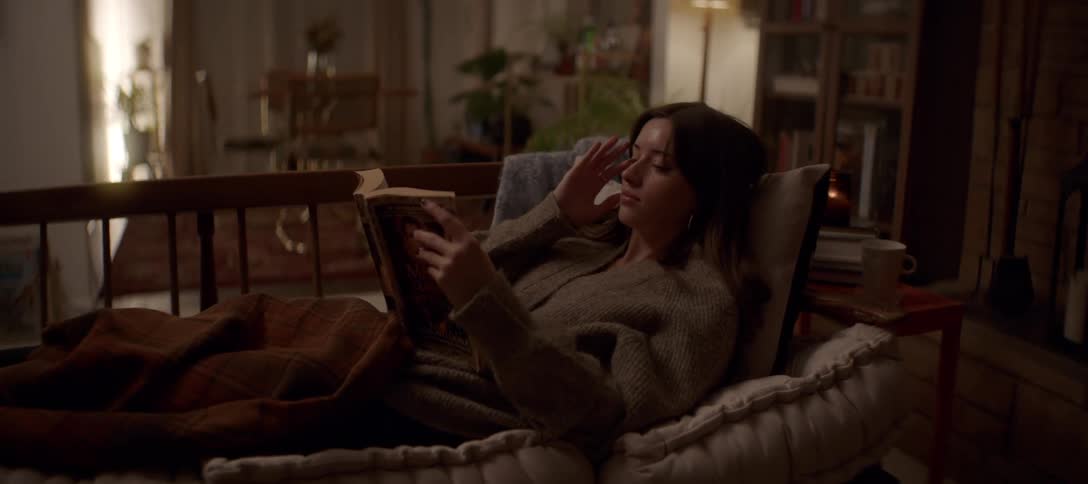 A young woman relaxes on a cozy couch in a warmly lit living room at night, engrossed in reading a book
