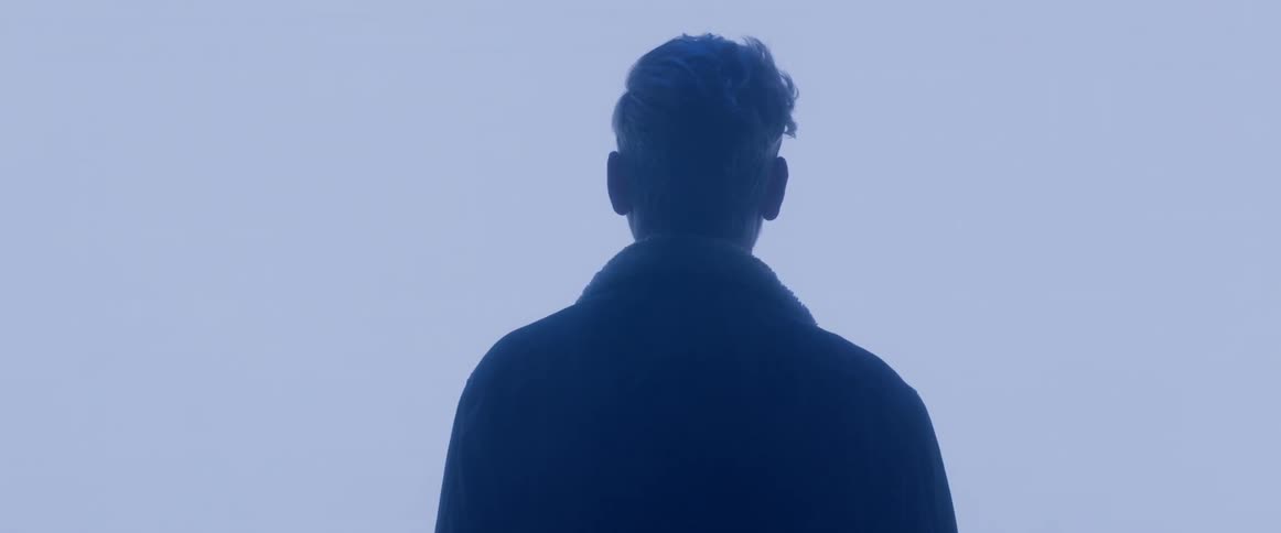 A man facing away from the camera wearing a coat standing in the fog
