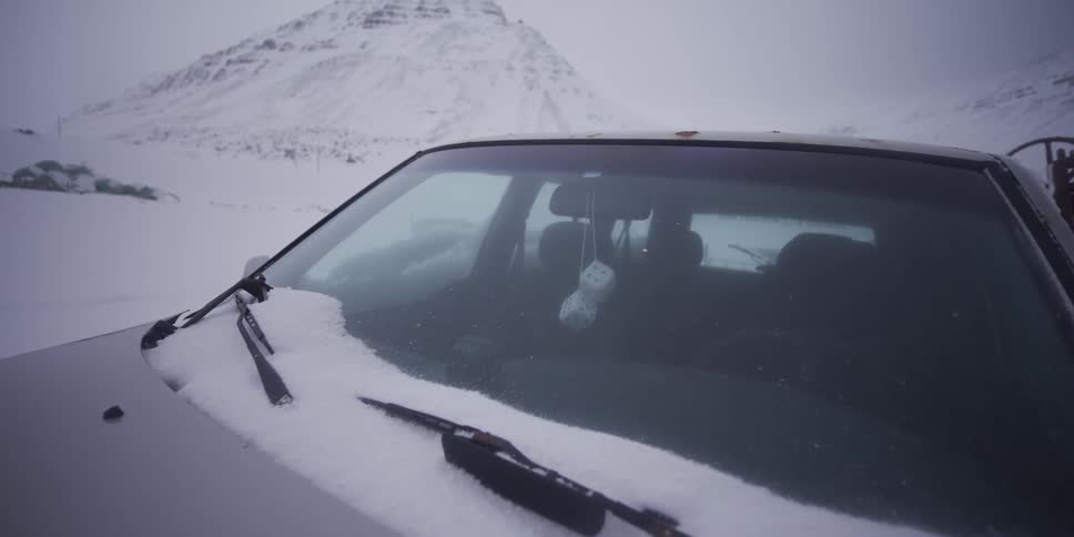A slow zoom into the windshield of a car covered in snow reveals fuzzy dice hanging from the mirror in a cold, mountainous landscape