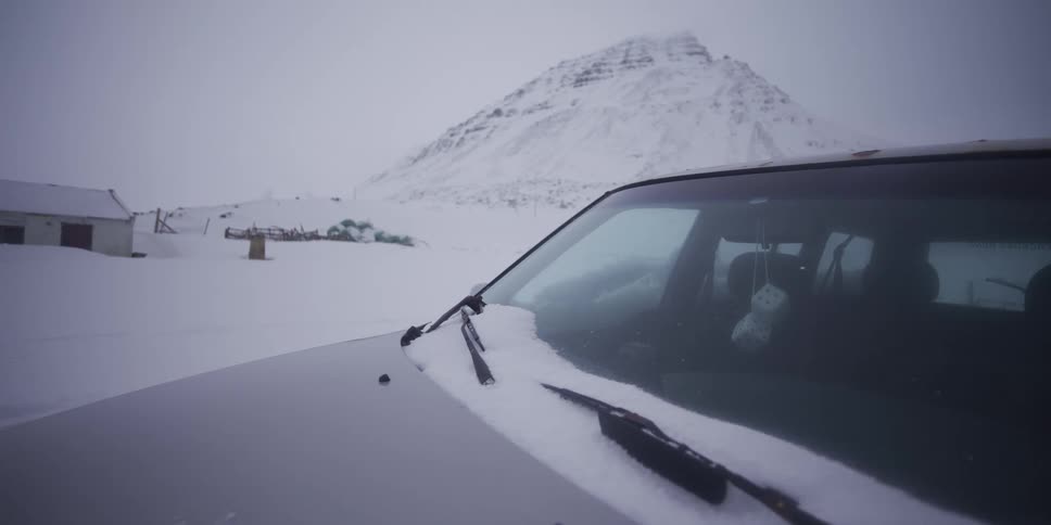 A close-up captures snowflakes landing on the windshield of a car parked in a remote, snowy landscape with mountains in the background