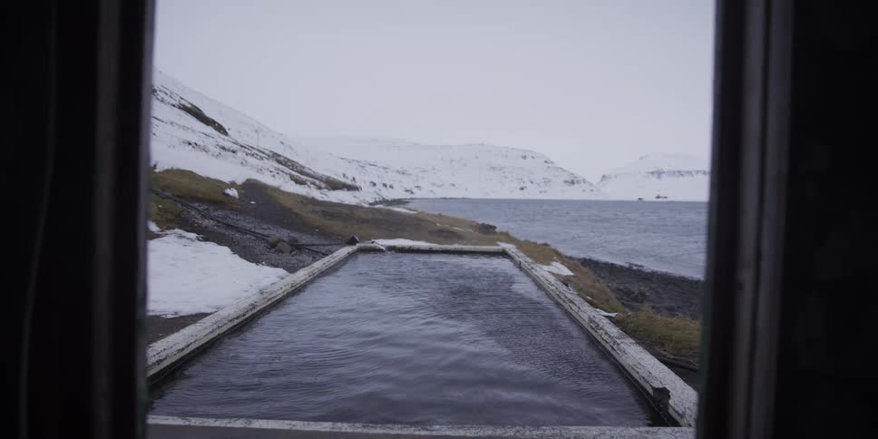 A slow-moving shot from a dark room reveals an outdoor geothermal hot spring overlooking a fjord on a snowy winter day