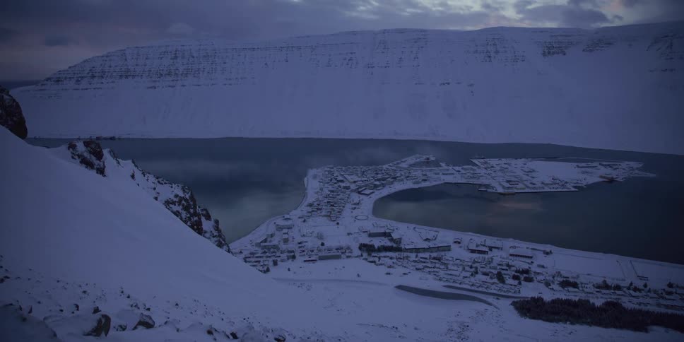 A scenic, slow tilt-up view from a snowy mountain reveals a remote coastal town nestled in a fjord during a cold winter twilight