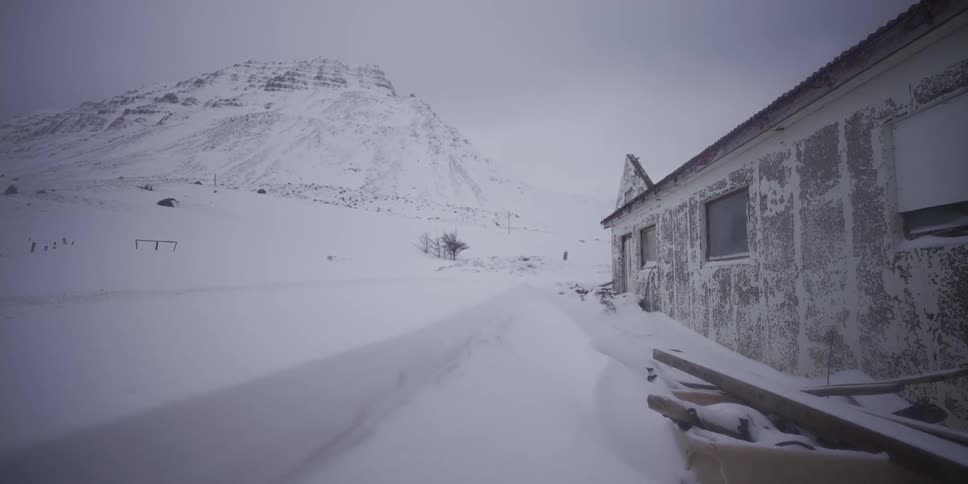 Howling winds whip snow across a desolate, frozen landscape next to a weathered, abandoned building at the base of a mountain