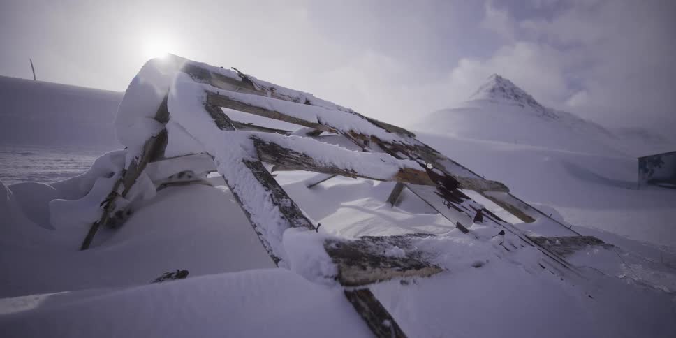 The weathered remains of a wooden structure are buried in deep snow in a desolate arctic landscape with mountains in the background
