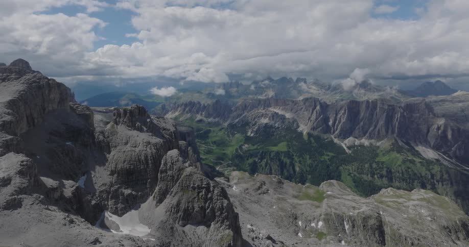 A stunning, sweeping aerial shot pushes over the rugged, rocky peaks of a mountain range, revealing a lush green valley below under a partly cloudy sky