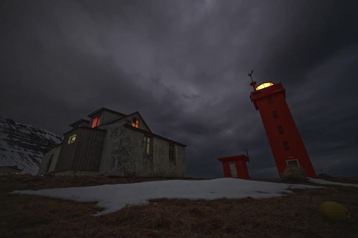 A bright red lighthouse flashes its beacon next to a weathered home at night while a person with a headlamp creates light trails on the snowy landscape