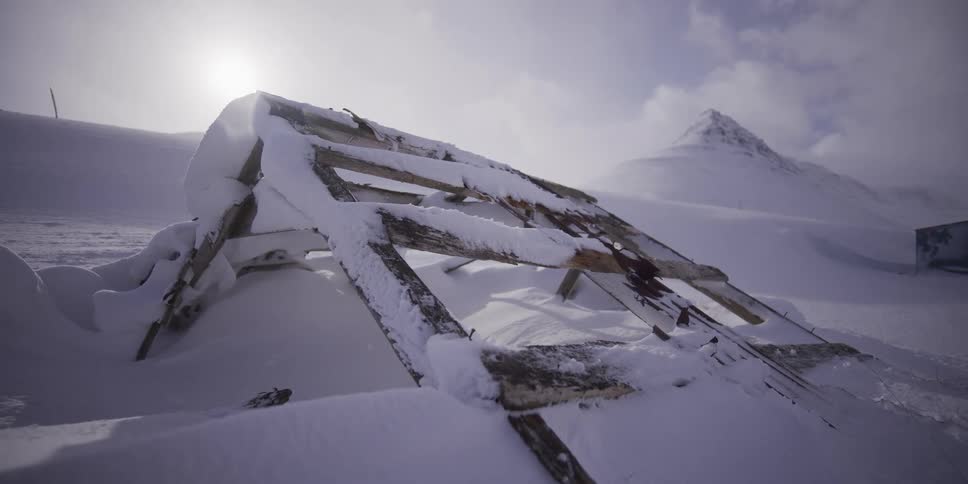 Slow zoom out from a snow-covered collapsed wooden building frame in a stark, frozen mountain wilderness