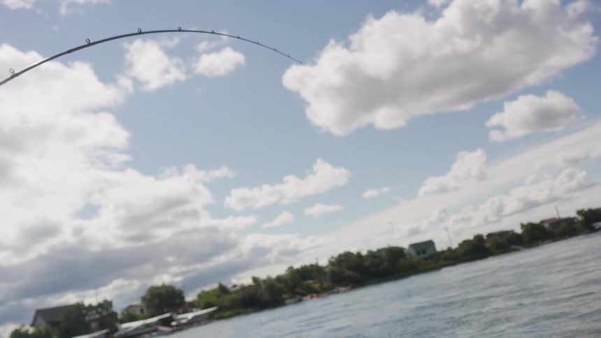 An angled shot shows a fishing rod bent under the weight of a fish against a bright blue sky with white clouds over the water