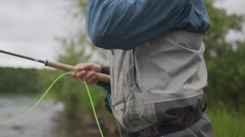 Close-up slow motion shot of a fly fisherman holding a rod and line while standing on a river bank