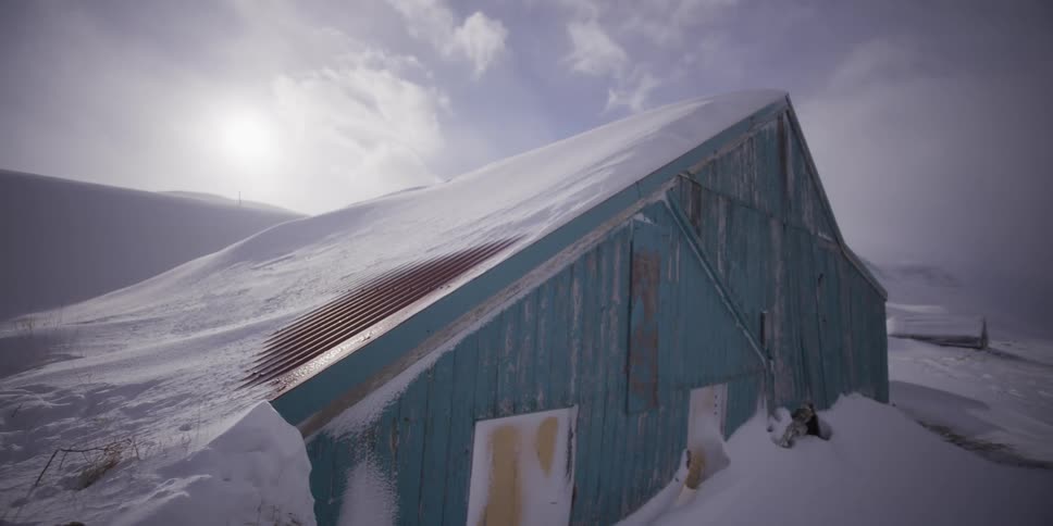 A weathered blue barn is slowly revealed from behind a deep snowdrift on a cold and windy winter day