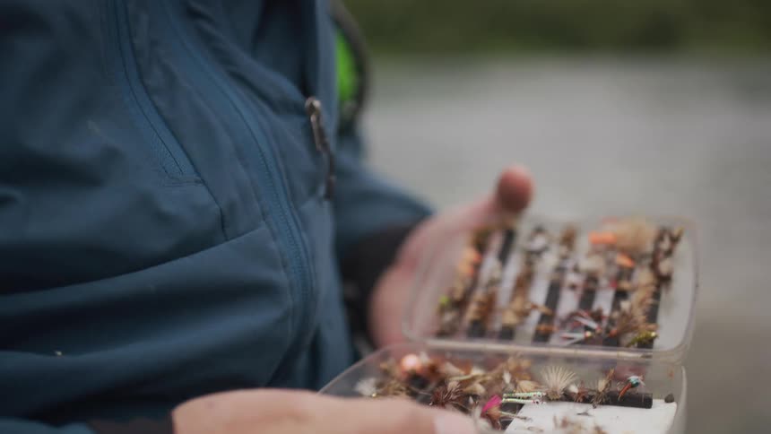 A fly fisherman slowly opens a clear tackle box to reveal a collection of colorful fishing flies and lures