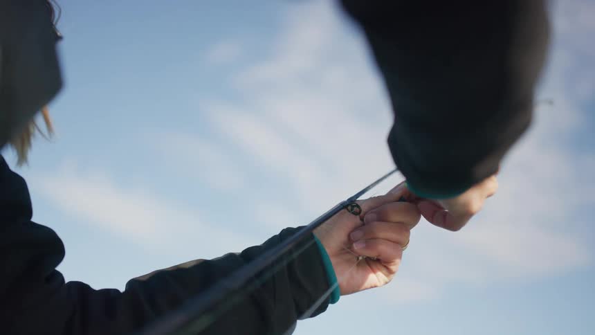 Slow motion close-up of hands preparing a fishing rod by attaching a bright green lure to the line against a blue sky