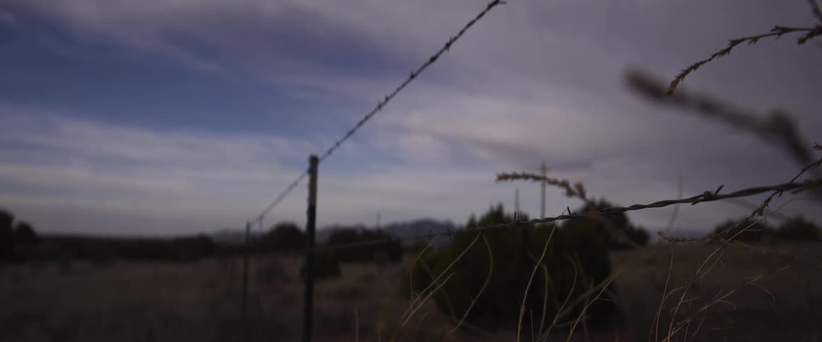 A slow rack focus shot across a barbed wire fence in a desolate rural landscape under a cloudy sky