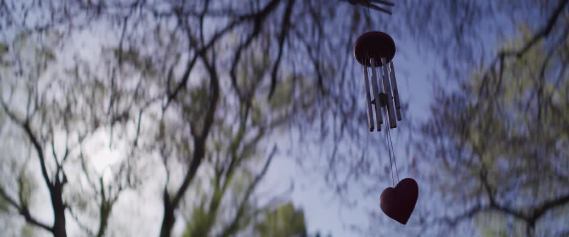A heart-shaped wind chime gently sways in the wind with soft-focus trees and a clear blue sky in the background