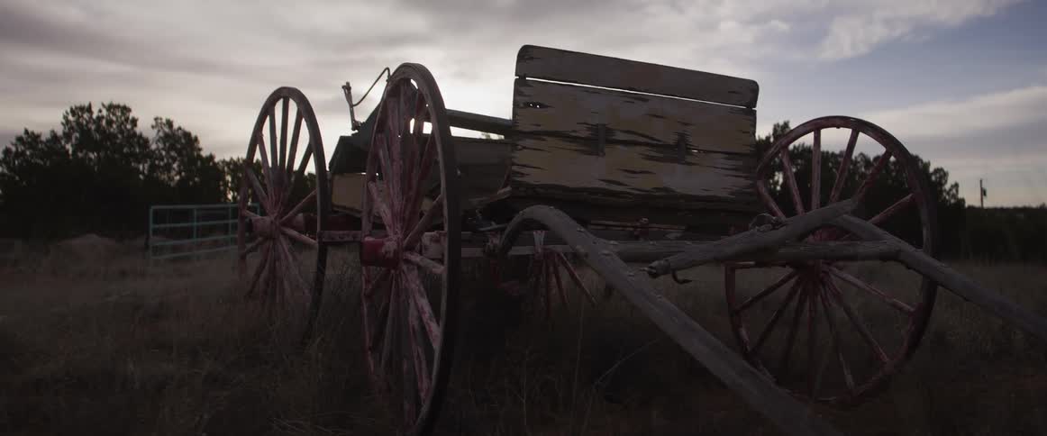 A slow dolly shot of an old, weathered pioneer wagon abandoned in a grassy field with the setting sun silhouetting its frame