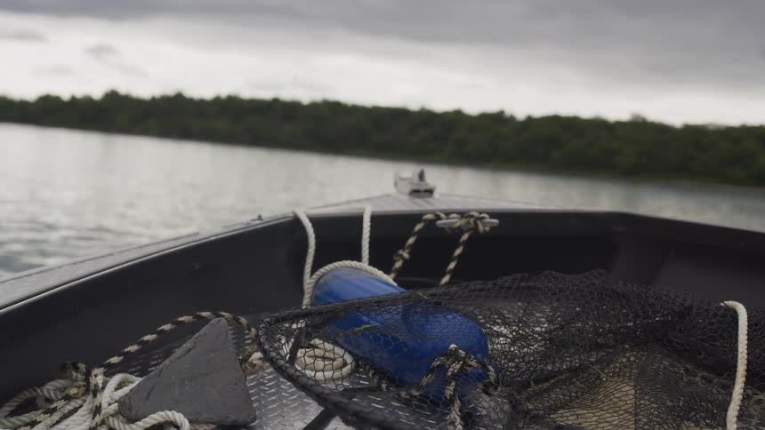 POV from a fishing boat with nets and buoys, moving across a calm lake on a cloudy day