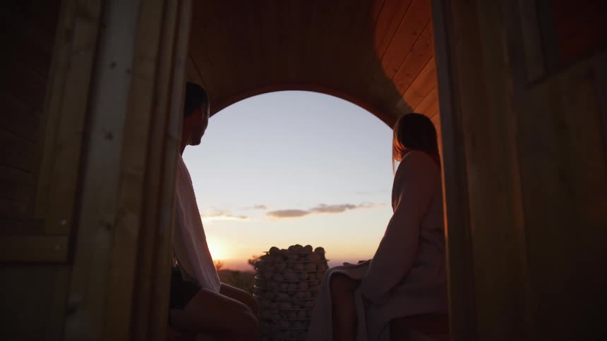 Slow pull-back shot reveals a silhouetted couple relaxing inside a cozy wooden barrel sauna, looking out at the serene sunset view