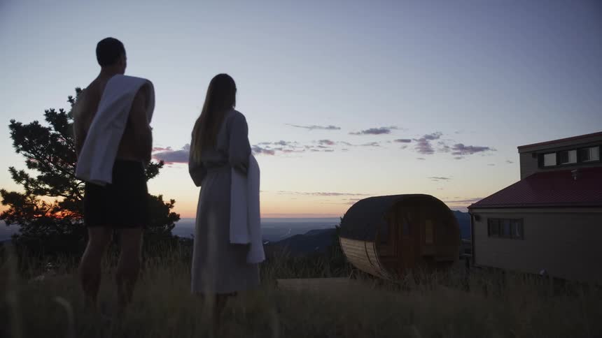 A serene couple stands on a grassy hillside overlooking a valley at sunset, ready for a relaxing session in their outdoor barrel sauna
