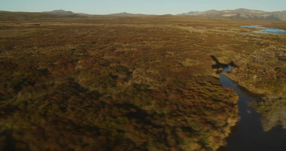 An aerial shot follows the shadow of a small bush plane as it flies over a remote river and vast autumn tundra
