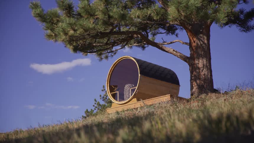 A unique wooden barrel sauna with a large circular window rests on a grassy hill beneath a large pine tree
