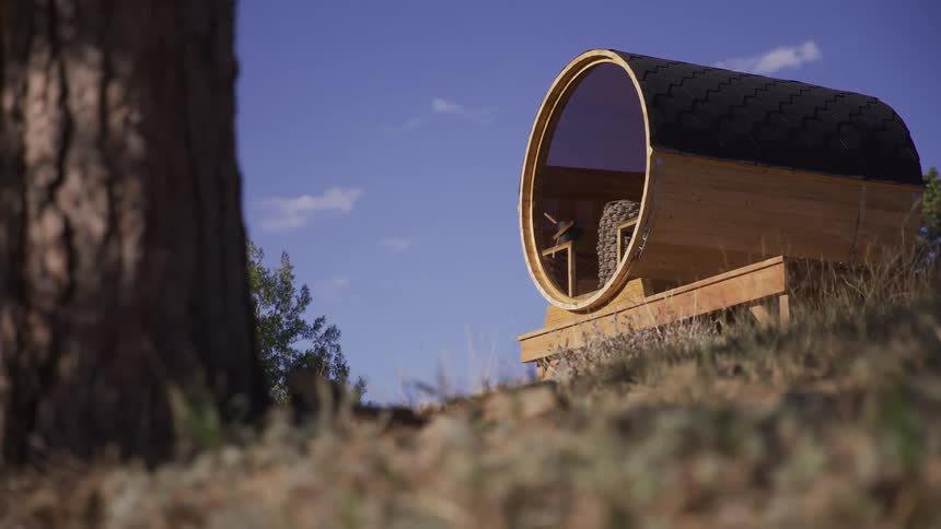 Low-angle dolly shot reveals a unique wooden barrel sauna with a panoramic window nestled on a tranquil hillside