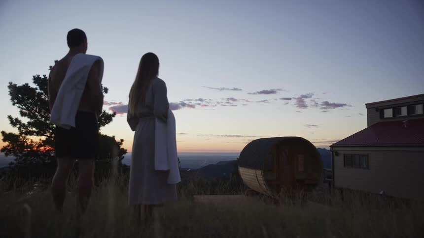 A couple in robes watches the sunset from a hilltop next to a modern home with a barrel sauna