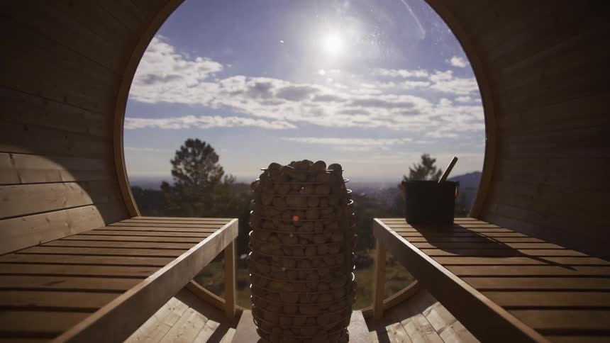 A tranquil push-in shot from inside a modern barrel sauna reveals a stunning panoramic view of a sunny landscape