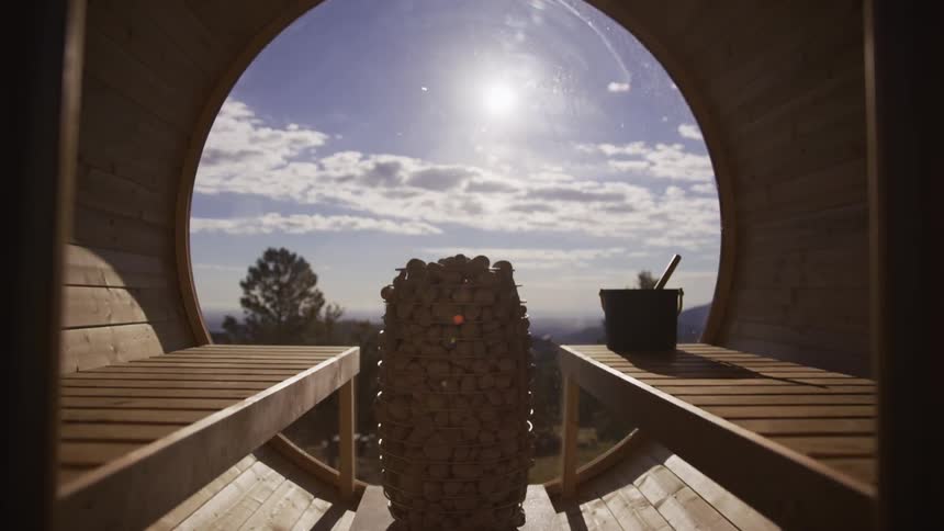 A slow dolly-out shot from inside a tranquil barrel sauna reveals a picturesque sunny landscape through a large panoramic window