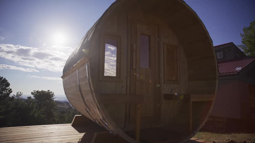 Slow dolly shot reveals a modern wooden barrel sauna on a deck under the bright sun