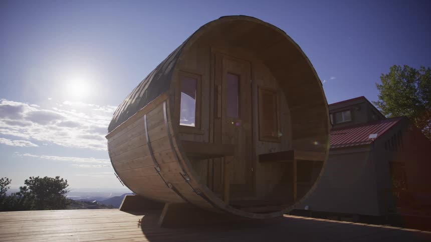 A unique barrel-shaped wooden sauna sits on an outdoor deck under the bright sun and a clear blue sky